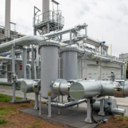 Kompleksowa instalacja przemysłowa z rurami, zbiornikami i konstrukcjami stalowymi na zewnątrz budynku. Widoczna wieża wyciągowa w tle.