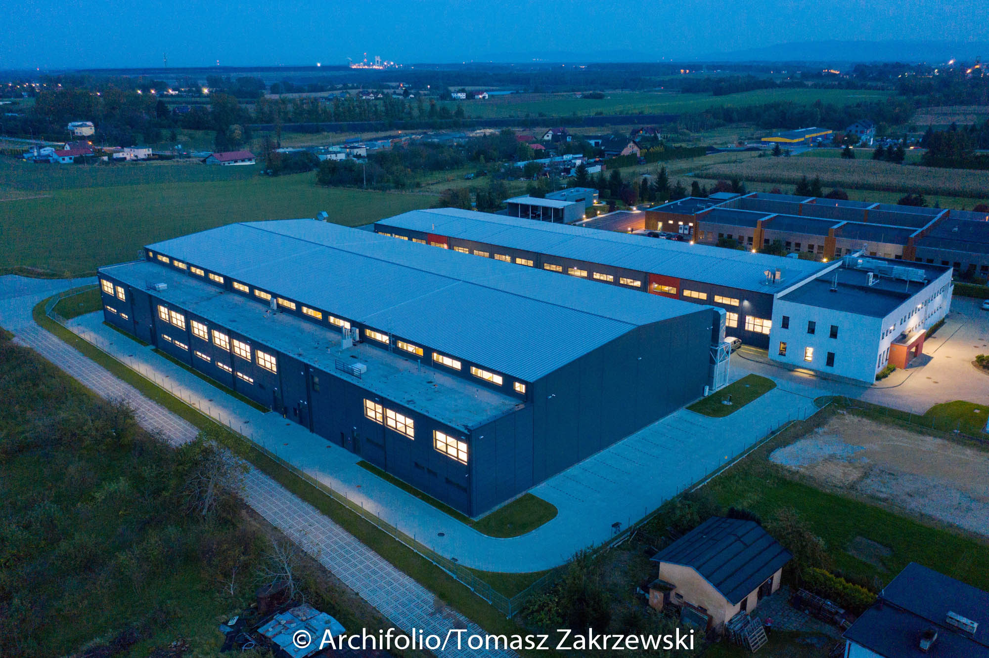 Wieczorny widok z lotu ptaka na nowoczesną halę przemysłową z oświetlonymi oknami, otoczoną zielenią i innymi budynkami. W tle widoczne są światła miasta.