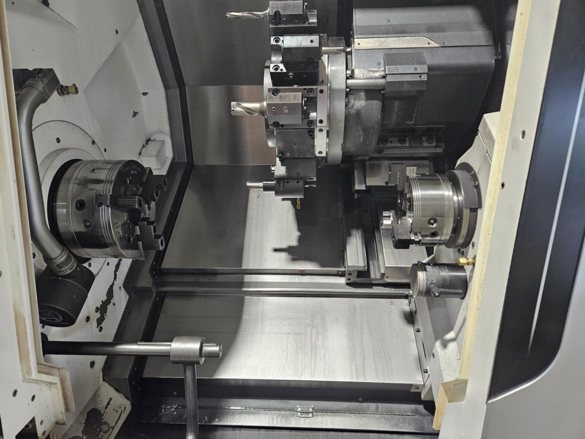 Wnętrze tokarki CNC z uchwytami, narzędziami i metalową powierzchnią roboczą, oświetlone światłem górnym. Widok z góry na precyzyjną maszynę.