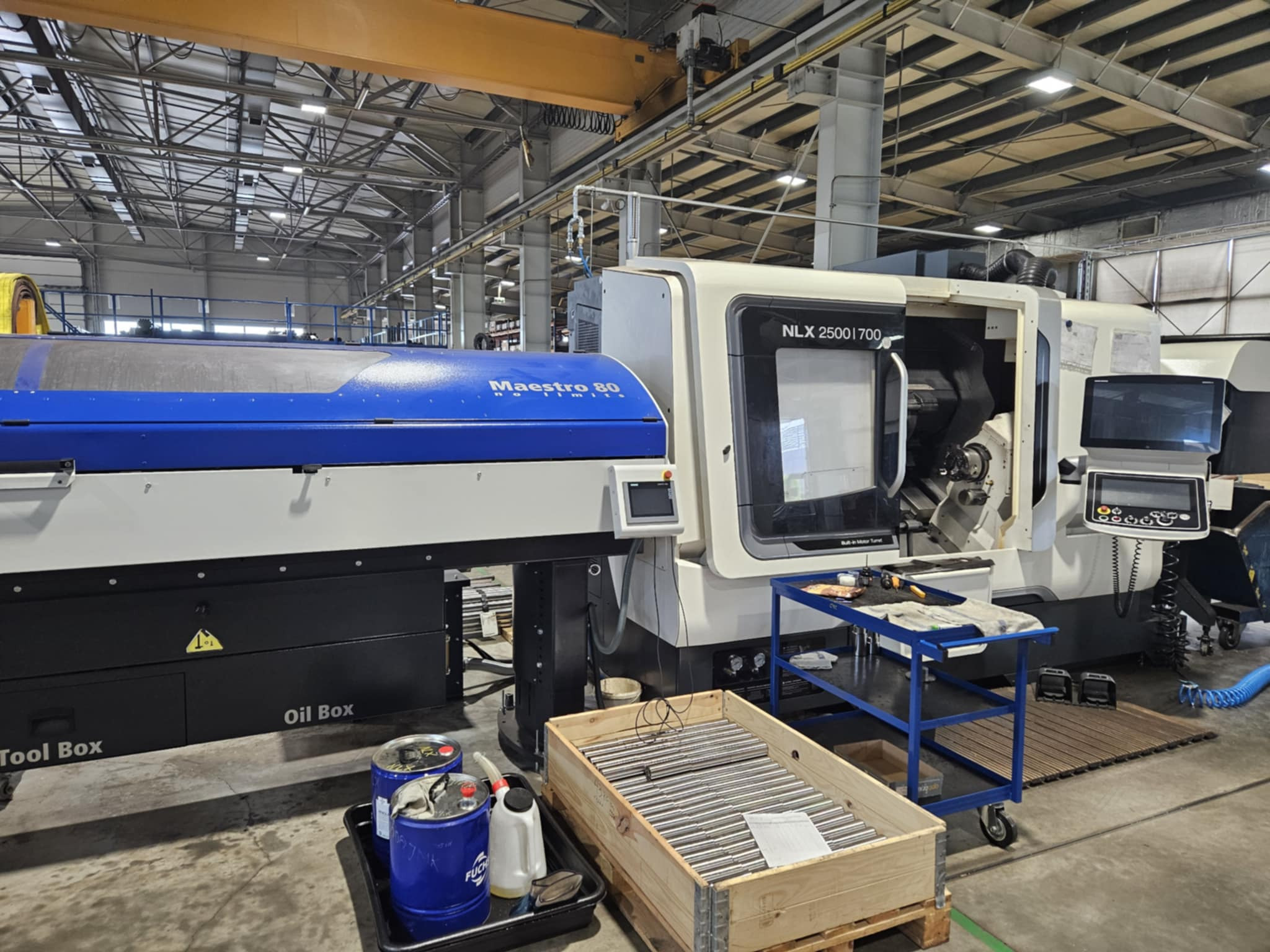 Nowoczesna tokarka CNC NLX 2500|700 z niebieską obudową Maestro 80 w hali produkcyjnej, z widocznymi narzędziami i elementami wyposażenia, jak skrzynka narzędziowa i wózek.