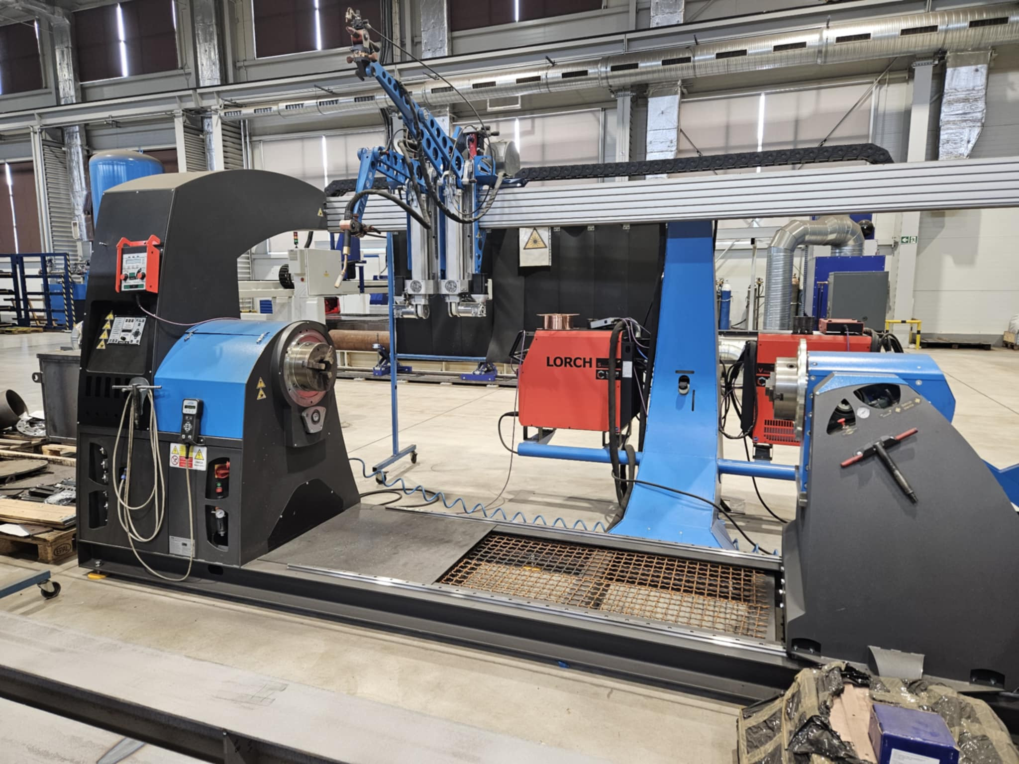 Automatyczne stanowisko spawalnicze z niebieskim robotem, w hali produkcyjnej. Widoczne elementy sterujące i osłony bezpieczeństwa. Na podłodze metalowe elementy.