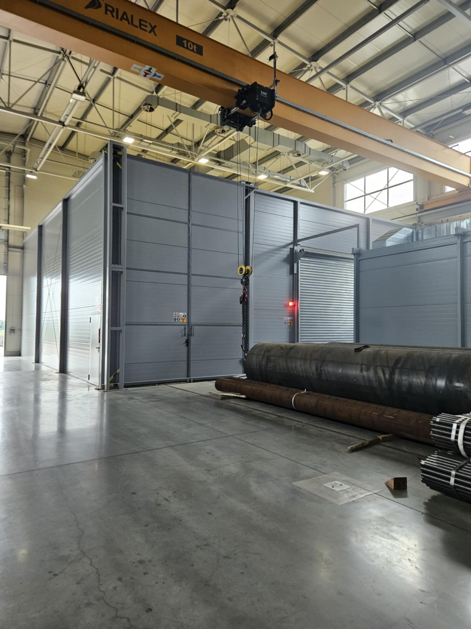 Wnętrze hali produkcyjnej z suwnicą i konstrukcjami stalowymi. Widoczne rury i zamknięta komora. Oświetlenie przemysłowe.