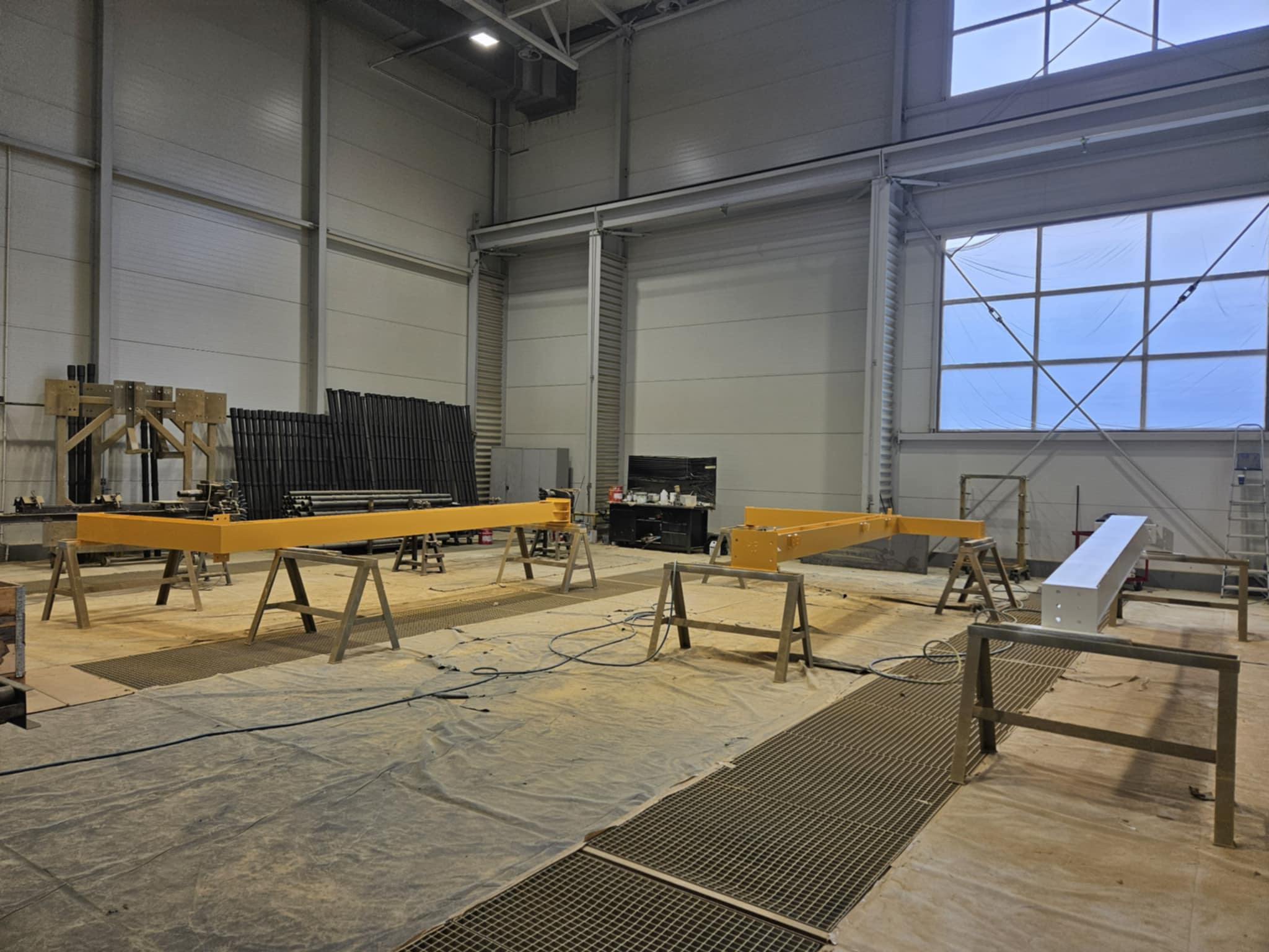 Wnętrze hali z żółtymi elementami konstrukcyjnymi na kozłach malarskich, przygotowanymi do dalszej obróbki. W tle widoczne rury i konstrukcja wsporcza.