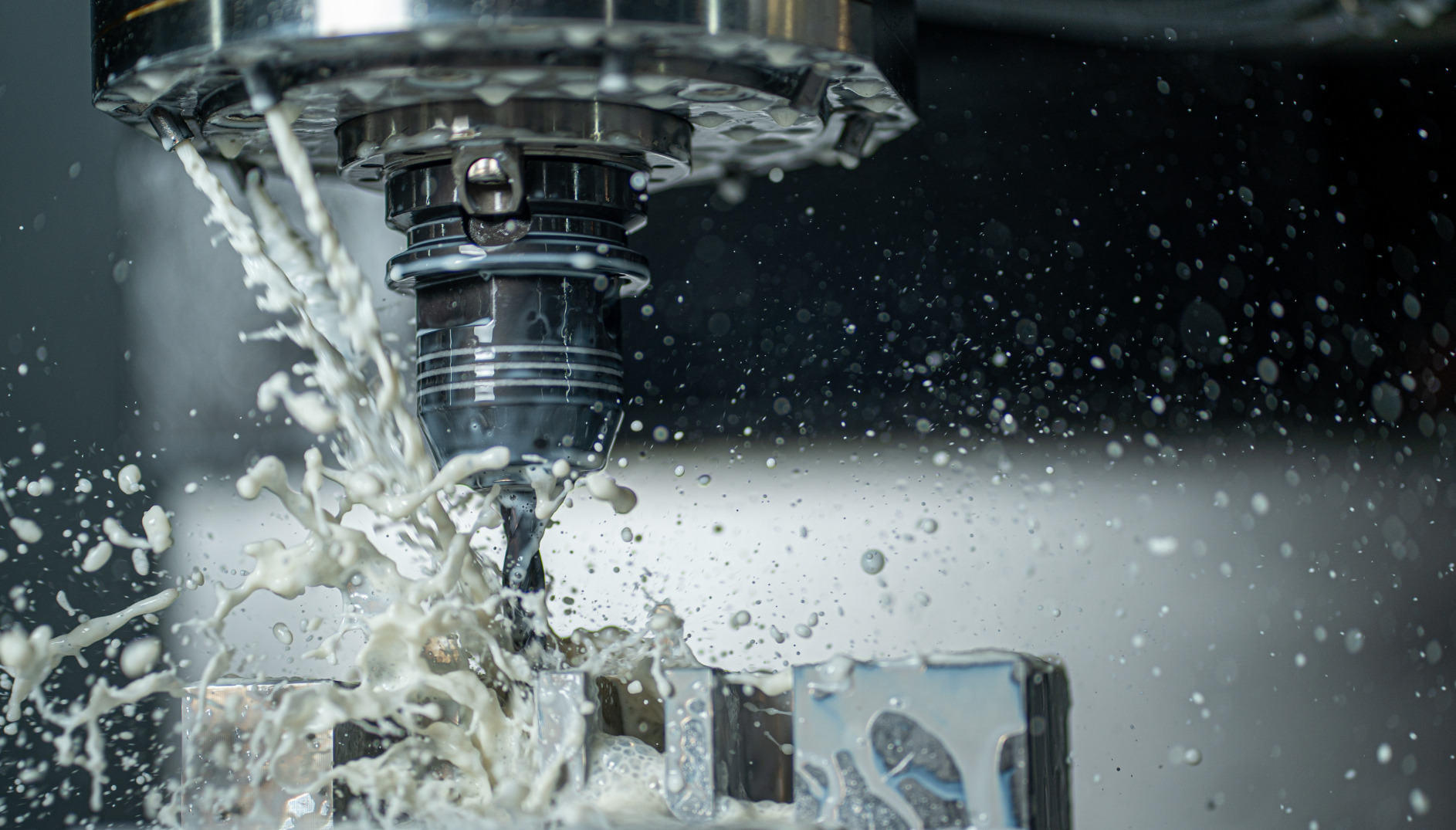 Precyzyjna obróbka metalu na maszynie CNC z chłodziwem, w zbliżeniu. Wióry i płyn chłodzący w ruchu, metalowy element obrabiany. Szare tło.