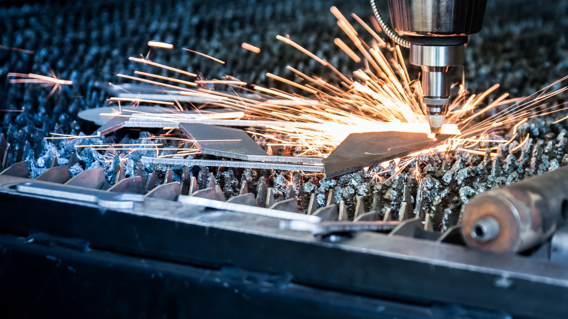 Precyzyjne cięcie metalu laserem, iskry w locie, fragmenty obrobionego materiału na perforowanym stole. Detal maszyny w tle, chłodna kolorystyka.