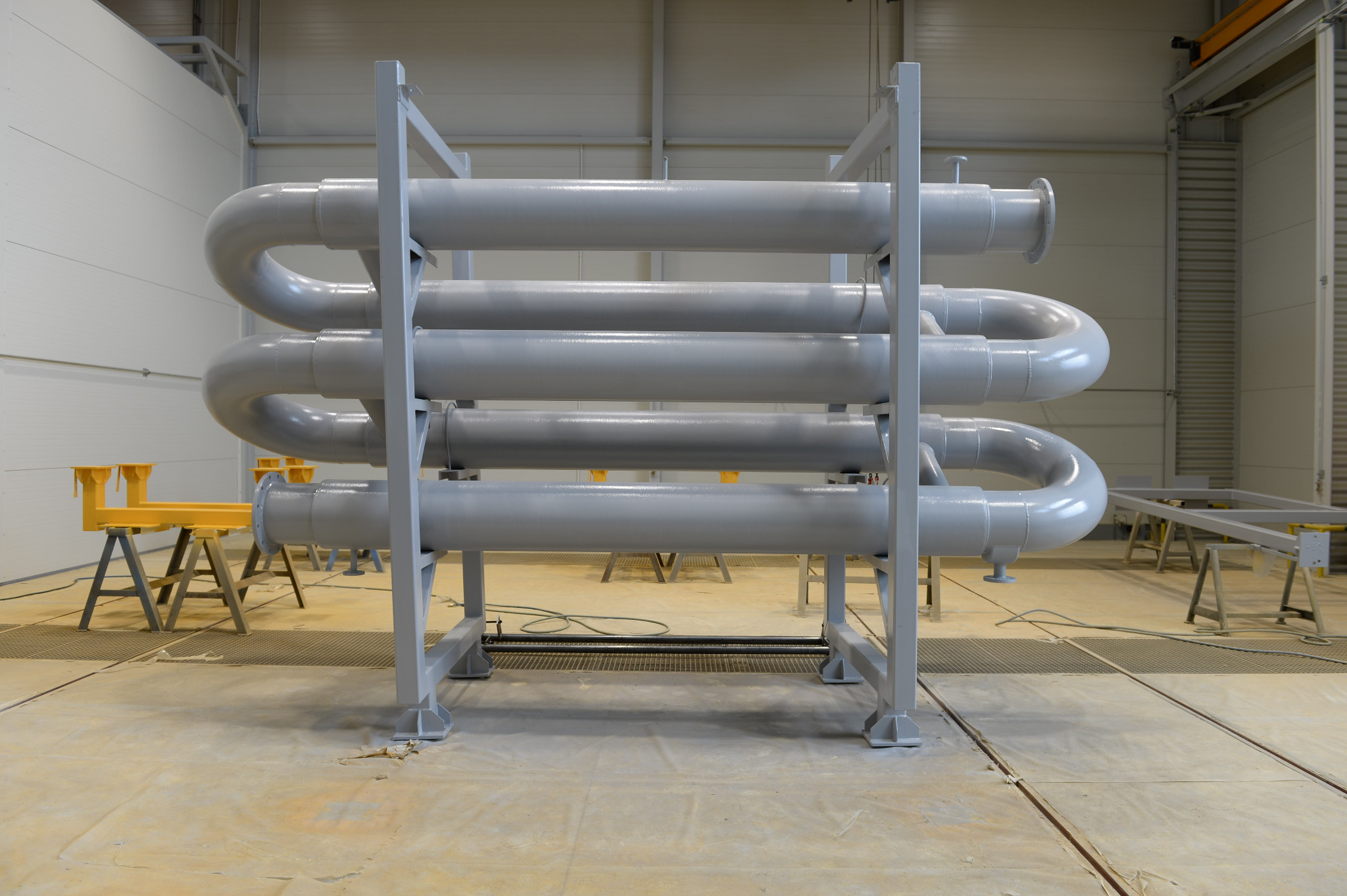 Szara, stalowa konstrukcja z rurami ułożonymi w spiralę, na tle hali produkcyjnej. Widoczne elementy wsporcze i podesty malarskie. Charakterystyczne połączenia kołnierzowe.