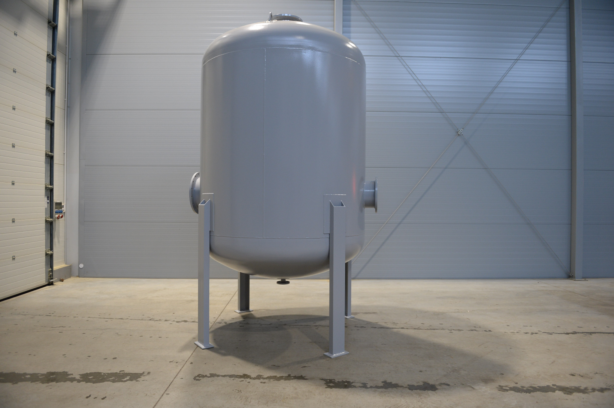 Szary, pionowy zbiornik ciśnieniowy na czterech nogach, z widocznymi króćcami przyłączeniowymi, na tle szarej ściany hali.
