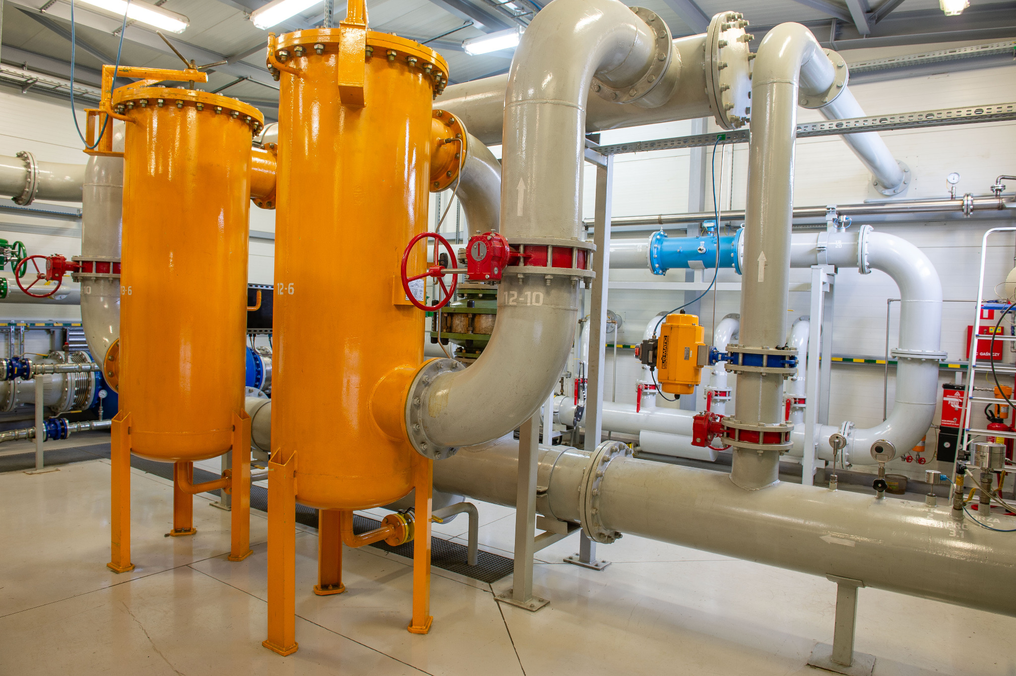 Instalacja przemysłowa z żółtymi zbiornikami i szarymi rurami, widoczne zawory z czerwoną armaturą. Jasne oświetlenie hali, białe ściany i podłoga.