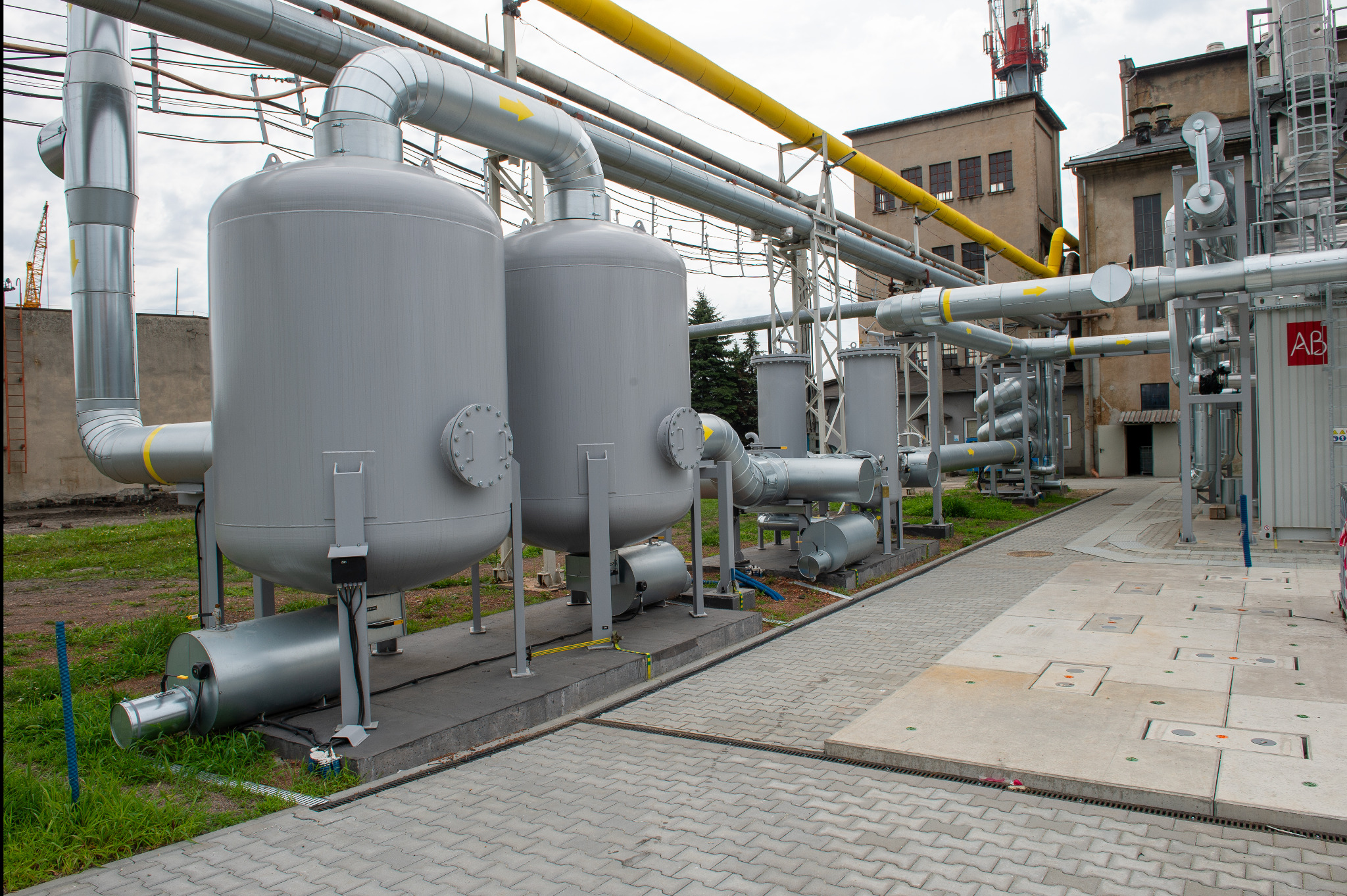 Kompleksowa instalacja przemysłowa z zbiornikami i rurociągami na zewnątrz budynku, widok z poziomu chodnika z kostki brukowej. Żółte oznaczenia na rurach.