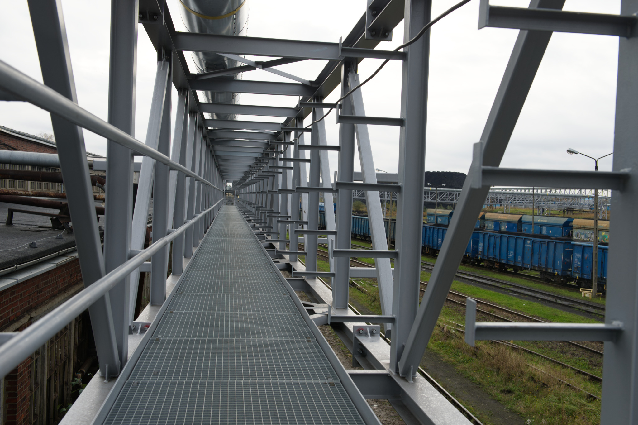 Długa, metalowa kładka przemysłowa z kratownicą, widok z perspektywy, z niebieskimi wagonami kolejowymi w tle. Konstrukcja pomalowana na szaro.