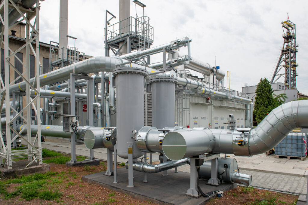 Kompleksowa instalacja przemysłowa z rurami, zbiornikami i konstrukcjami stalowymi na zewnątrz budynku. Widoczna wieża wyciągowa w tle.