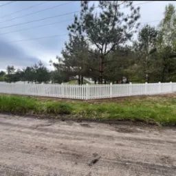 Białe, drewniane ogrodzenie sztachetowe z brązową listwą, otaczające posesję z zielenią, widziane z poziomu gruntu na tle drzew i pochmurnego nieba.