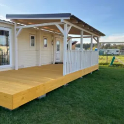 Żółty taras drewniany przy domu z białymi elementami konstrukcyjnymi i barierką. W tle plac zabaw i częściowo widoczna osoba montująca element.