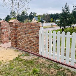 DzikWood - Nowoczesne ogrodzenie z białych sztachet i ceglanego murku, otaczające posesję z widokiem na dom i zieleń. Elementy wykończeniowe z czerwonej cegły.