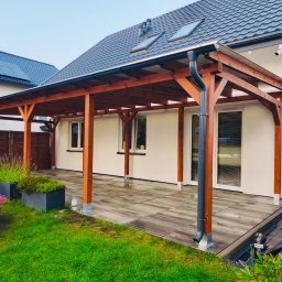 DzikWood - Zadaszony taras z drewnianą konstrukcją, wyłożony płytami. Widoczne donice z kwiatami i trawnik. Dom z szarą dachówką w tle.