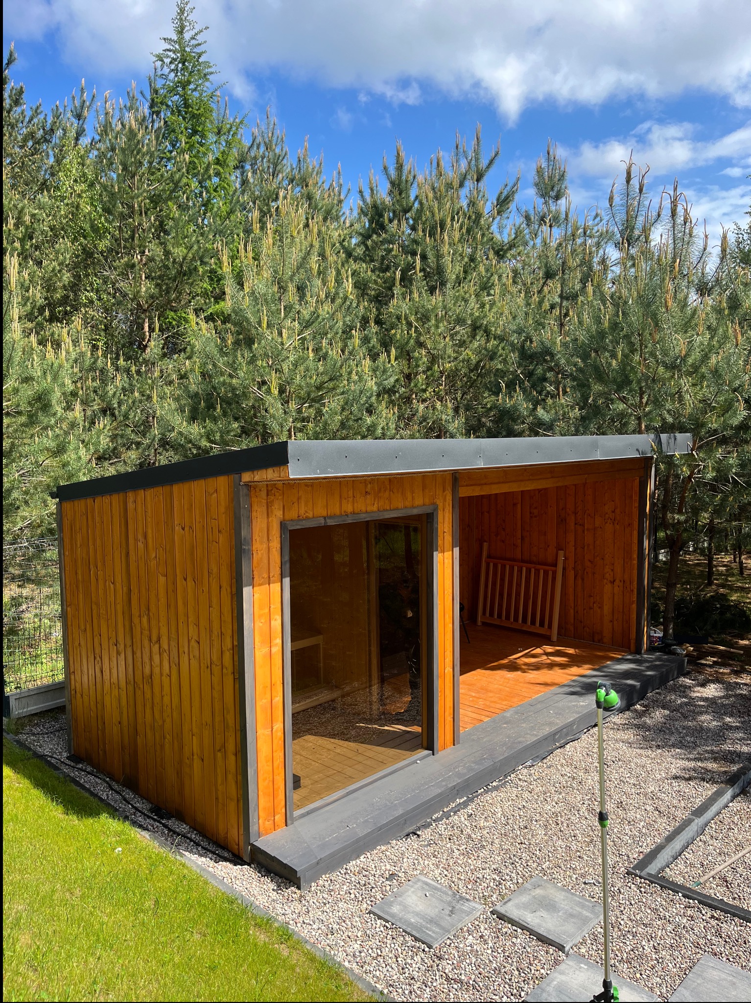 Nowoczesna, drewniana altana z przeszkloną ścianą i tarasem, otoczona zielenią. Widok z zewnątrz, w tle wysokie sosny i błękitne niebo.