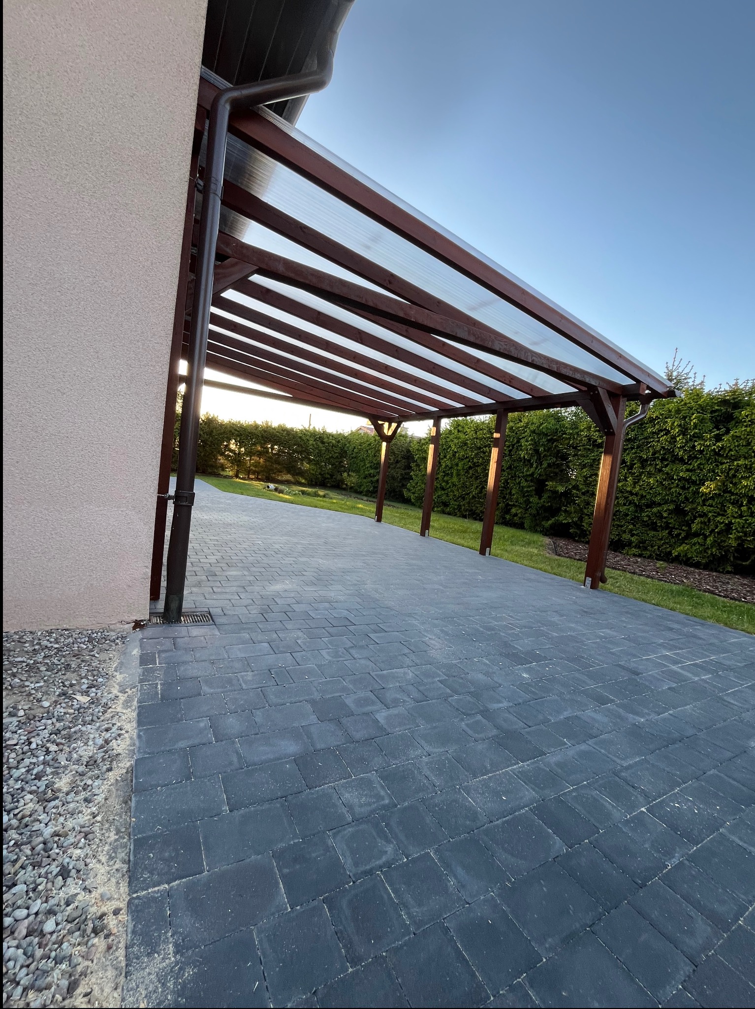 Zadaszenie tarasu z brązowego drewna i przezroczystego poliwęglanu, z widocznym systemem odprowadzania wody. Kostka brukowa prowadzi do zielonego ogrodu.