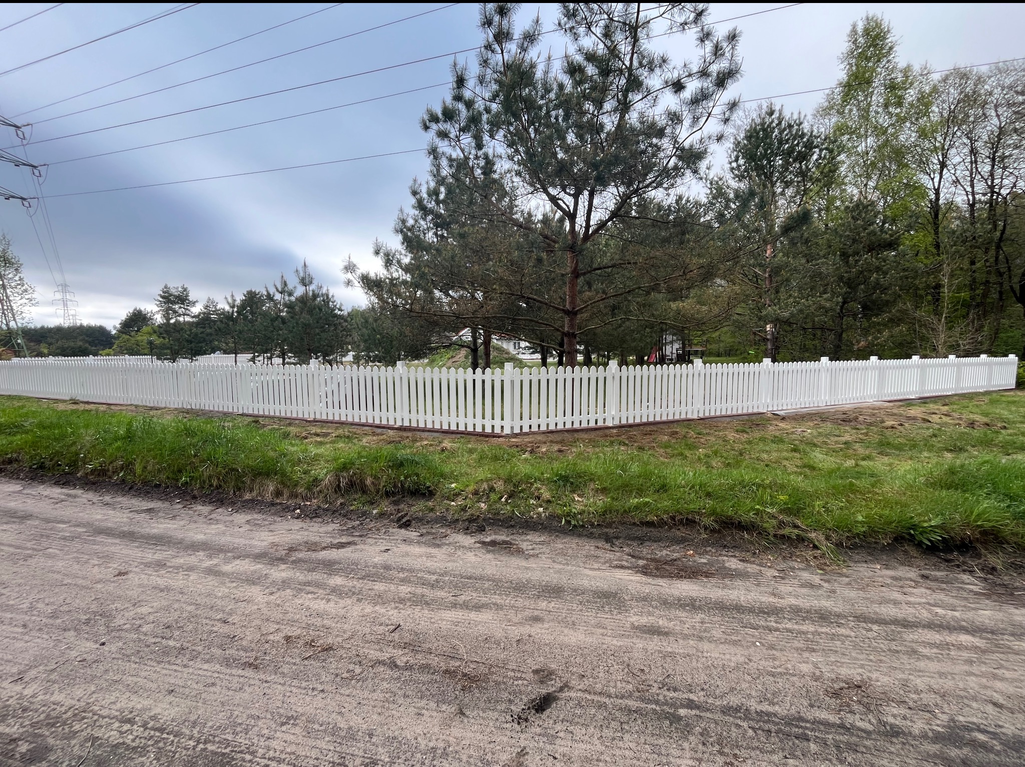 Białe, drewniane ogrodzenie sztachetowe z brązową listwą, otaczające posesję z zielenią, widziane z poziomu gruntu na tle drzew i pochmurnego nieba.