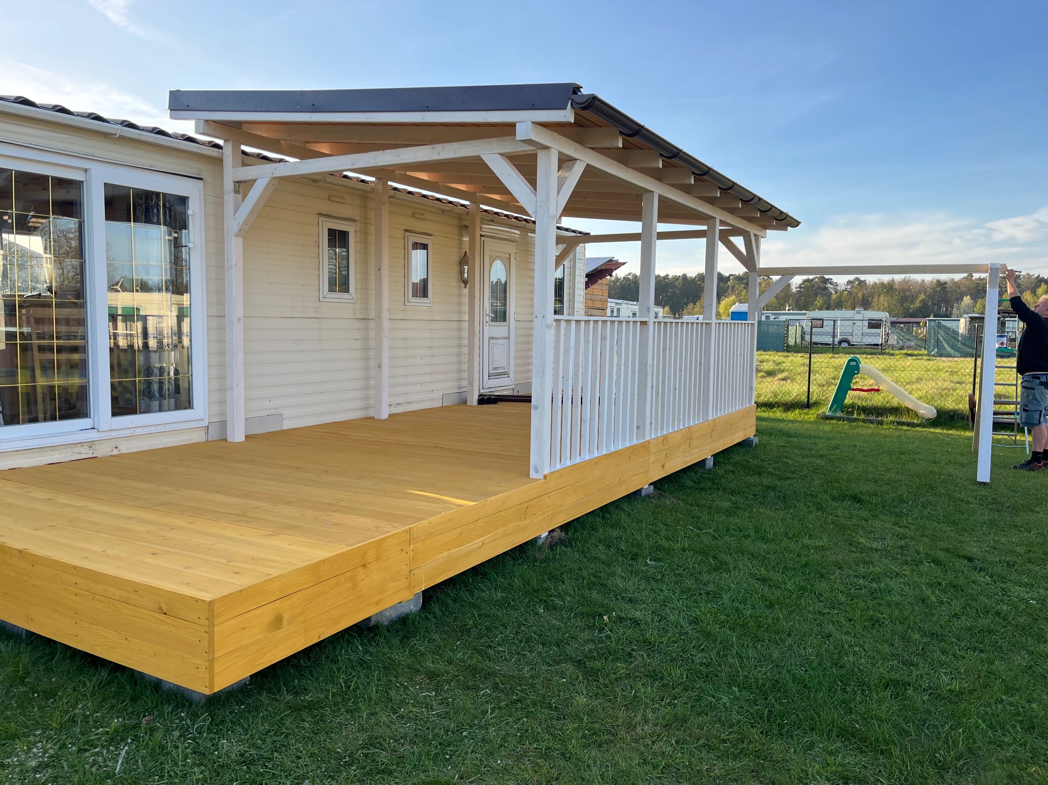 Żółty taras drewniany przy domu z białymi elementami konstrukcyjnymi i barierką. W tle plac zabaw i częściowo widoczna osoba montująca element.