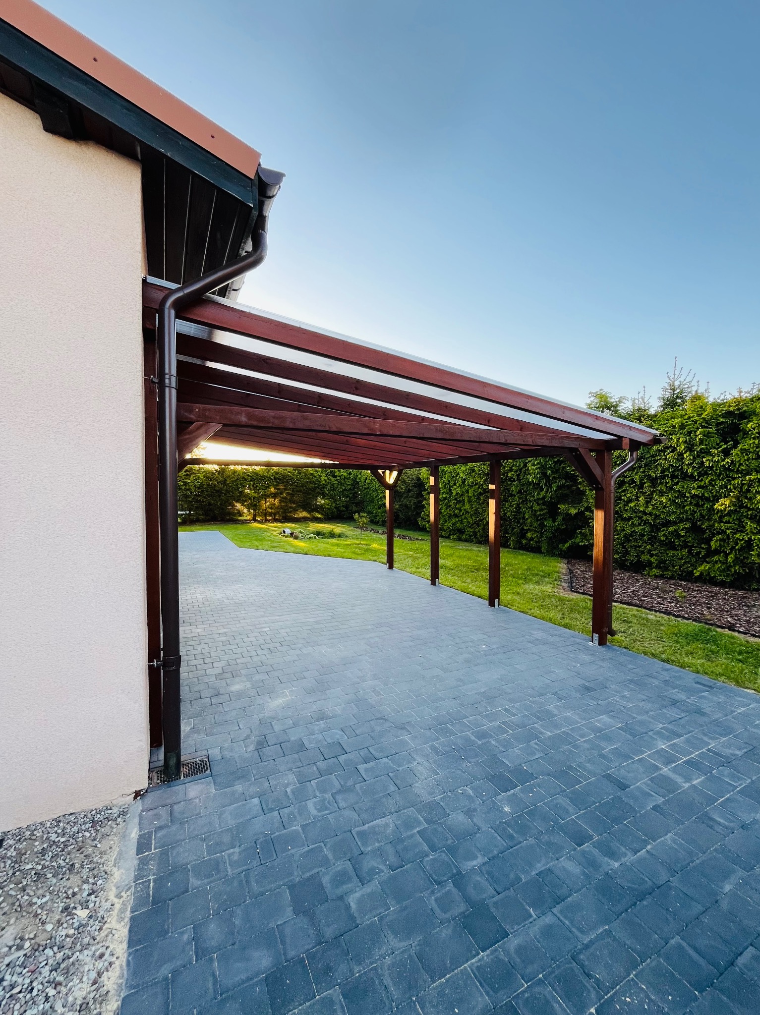 Ciemna, drewniana pergola z zadaszeniem, przylegająca do budynku z szarą kostką brukową. W tle zieleń ogrodu i błękitne niebo o zmierzchu.