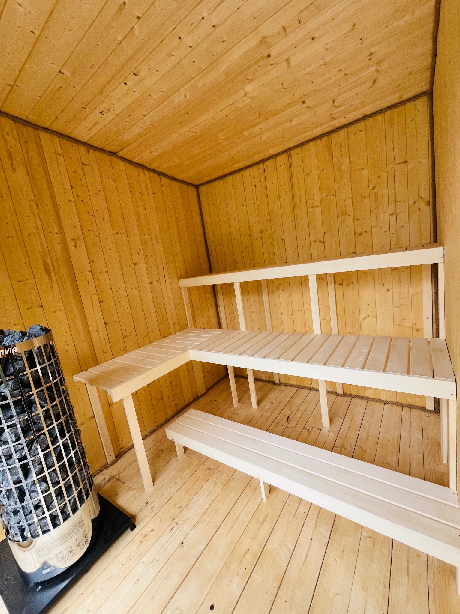 Wnętrze sauny z jasnego drewna: ławy, ściany i podłoga. Piec z kamieniami w rogu. Minimalistyczny design, surowe drewno, ciepłe oświetlenie.