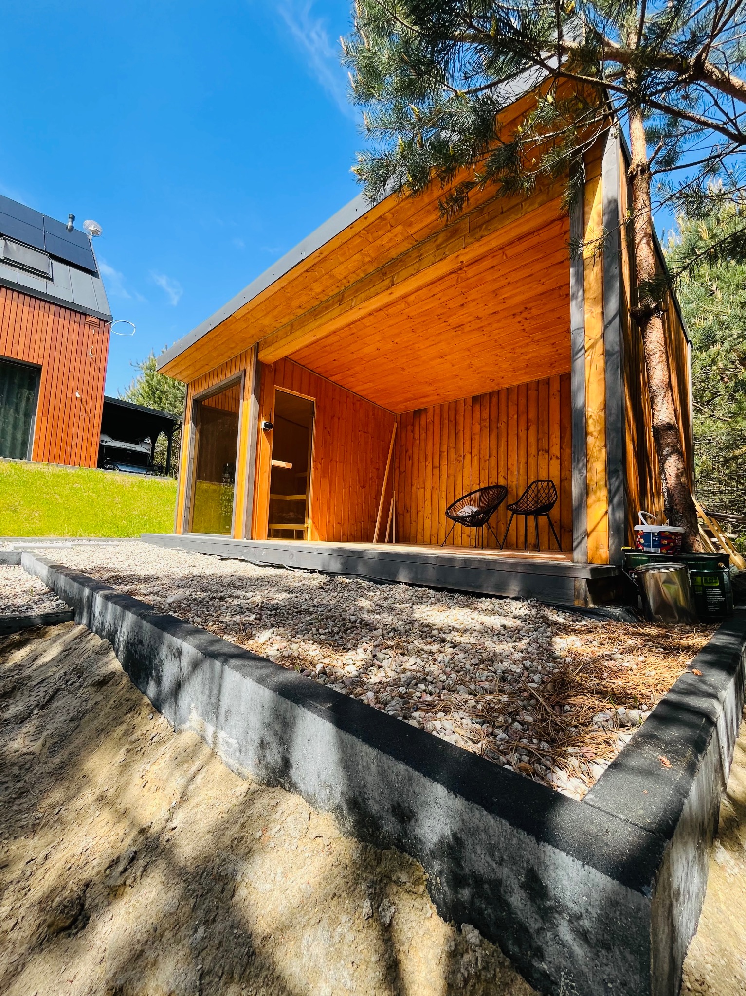 Drewniana altana z zadaszeniem i dwoma krzesłami na podeście, otoczona kamieniami i zielenią, w tle budynek z panelami słonecznymi i błękitne niebo.