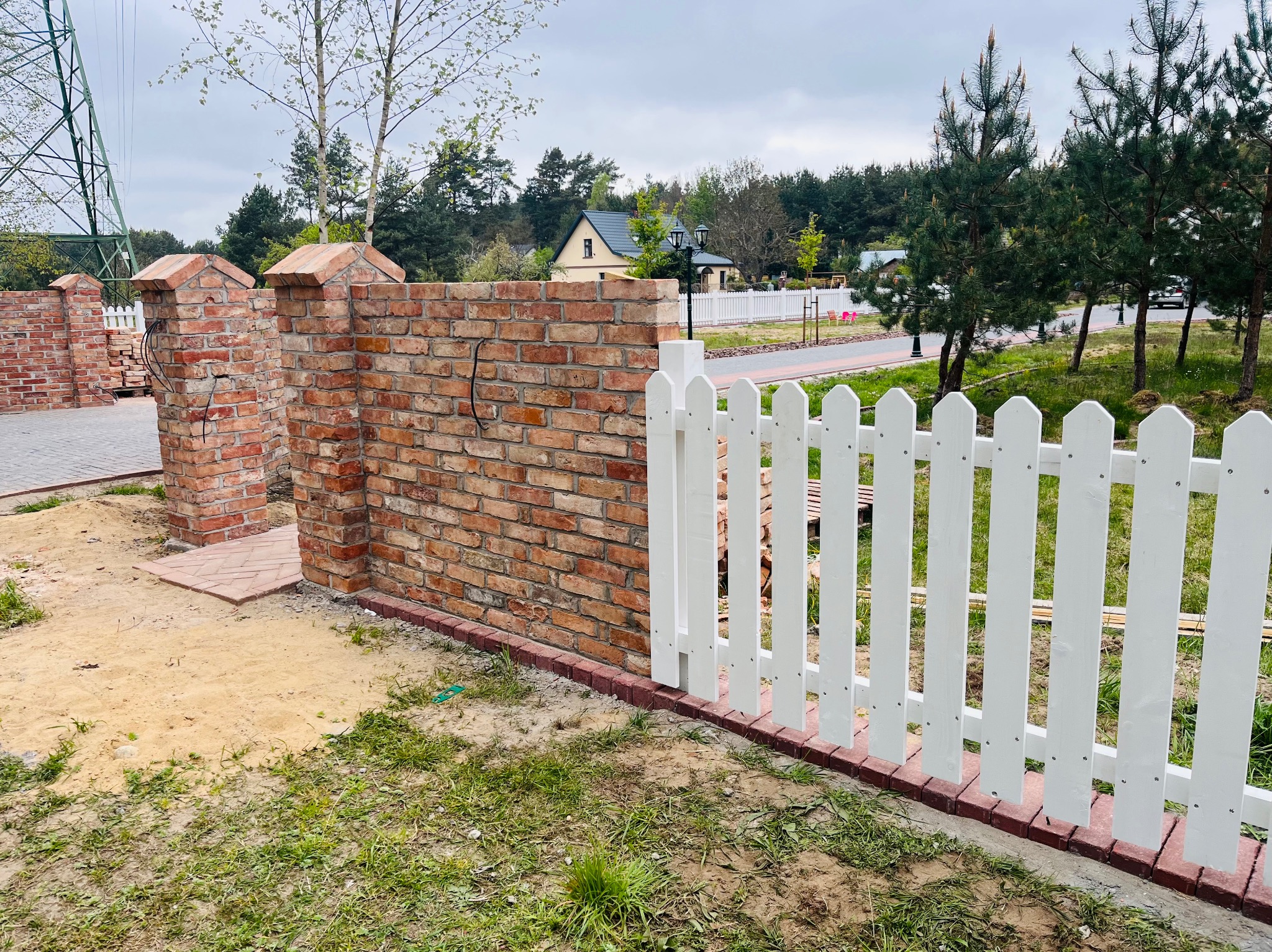 Nowoczesne ogrodzenie z białych sztachet i ceglanego murku, otaczające posesję z widokiem na dom i zieleń. Elementy wykończeniowe z czerwonej cegły.