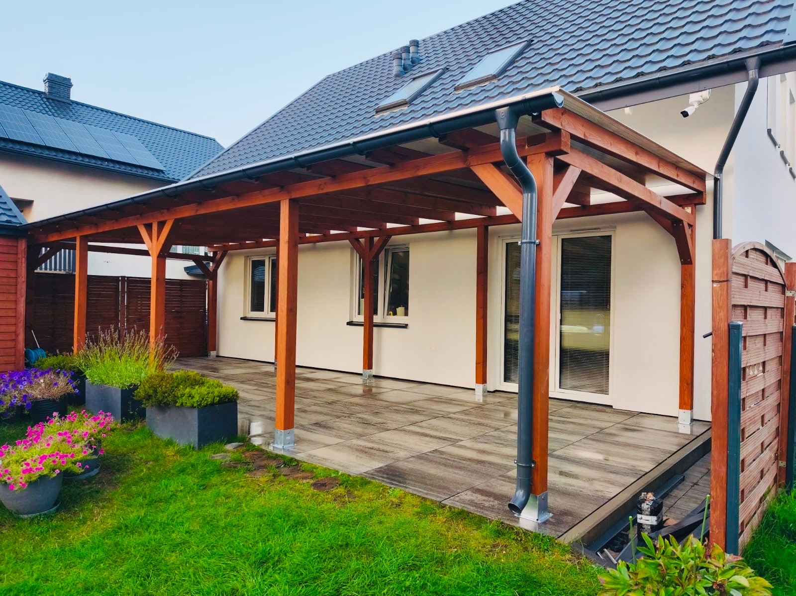 Zadaszony taras z drewnianą konstrukcją, wyłożony płytami. Widoczne donice z kwiatami i trawnik. Dom z szarą dachówką w tle.