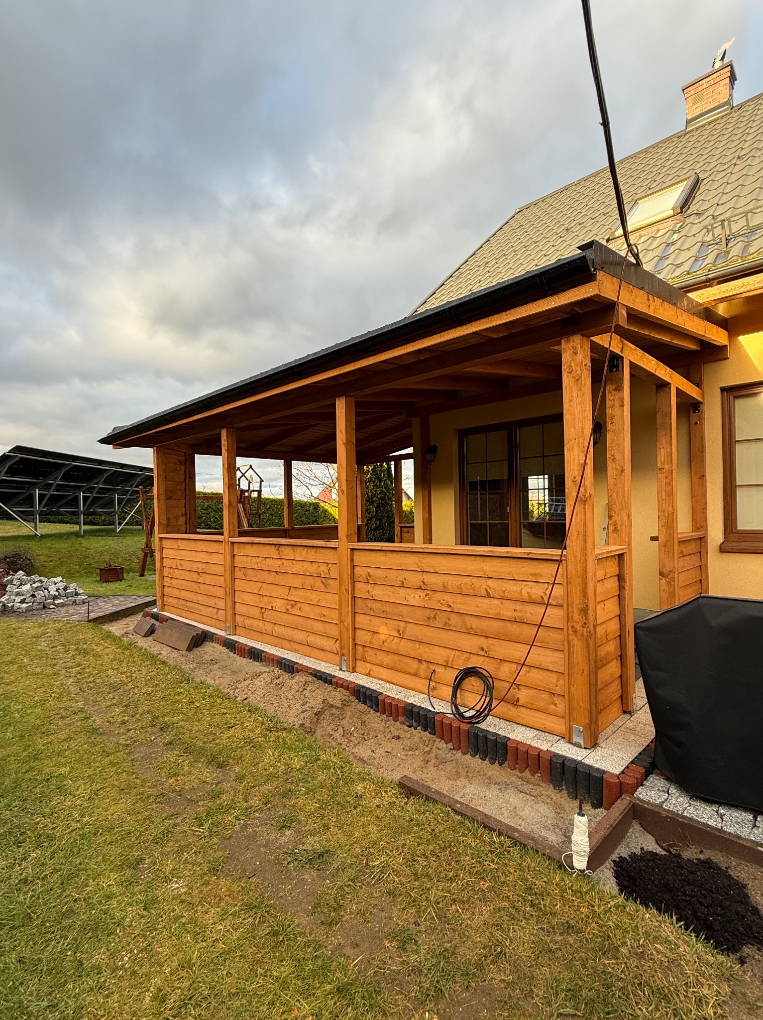 Drewniana, nowa altana przydomowa z płotkiem, widok z zewnątrz. Na trawniku leżą kostki brukowe. W tle dom z panelami słonecznymi i pochmurne niebo.