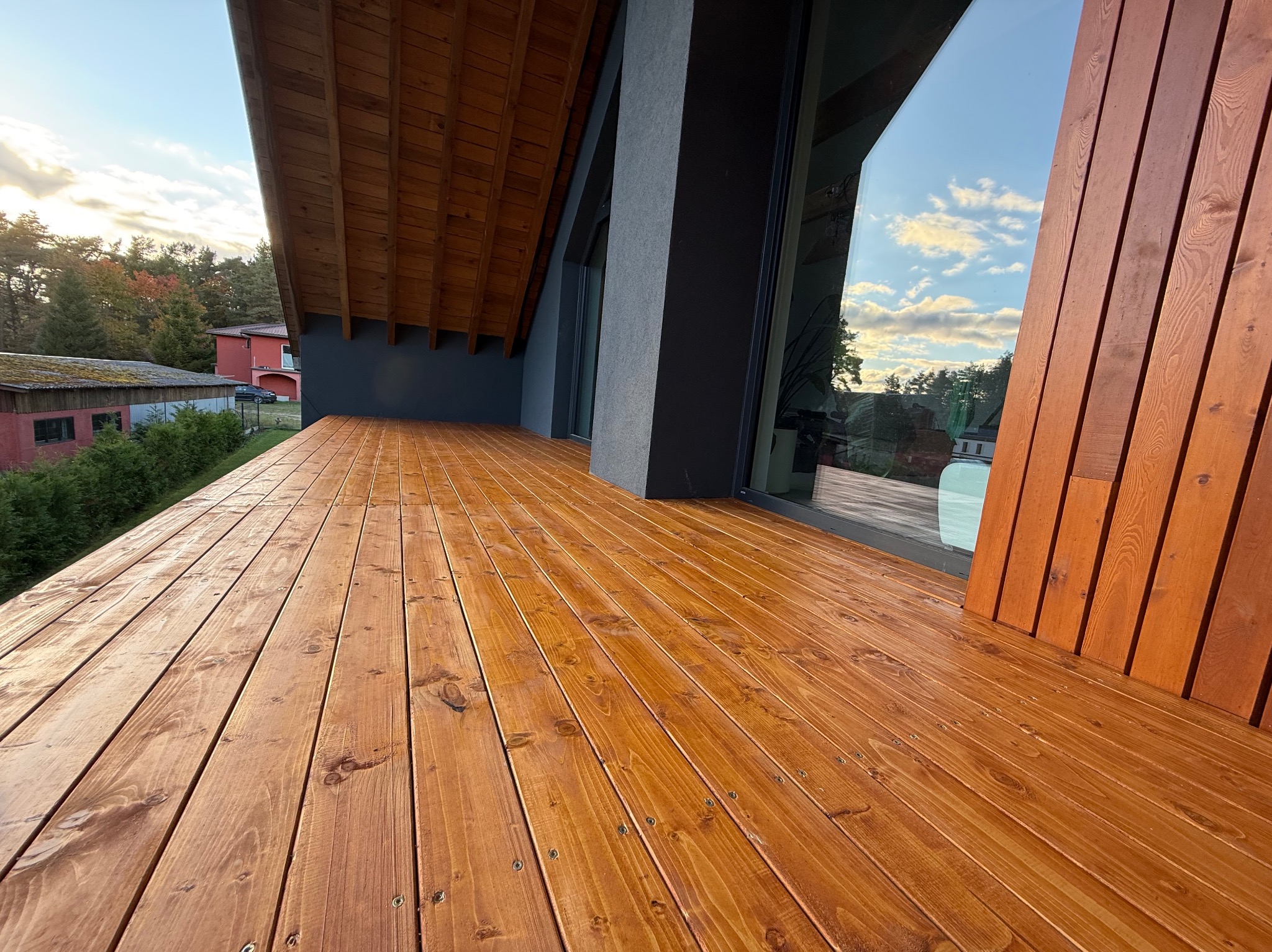 Błyszczący, drewniany taras przydomowy z widocznymi śrubami mocującymi deski, przylegający do nowoczesnego budynku z dużym oknem i drewnianą ścianą.