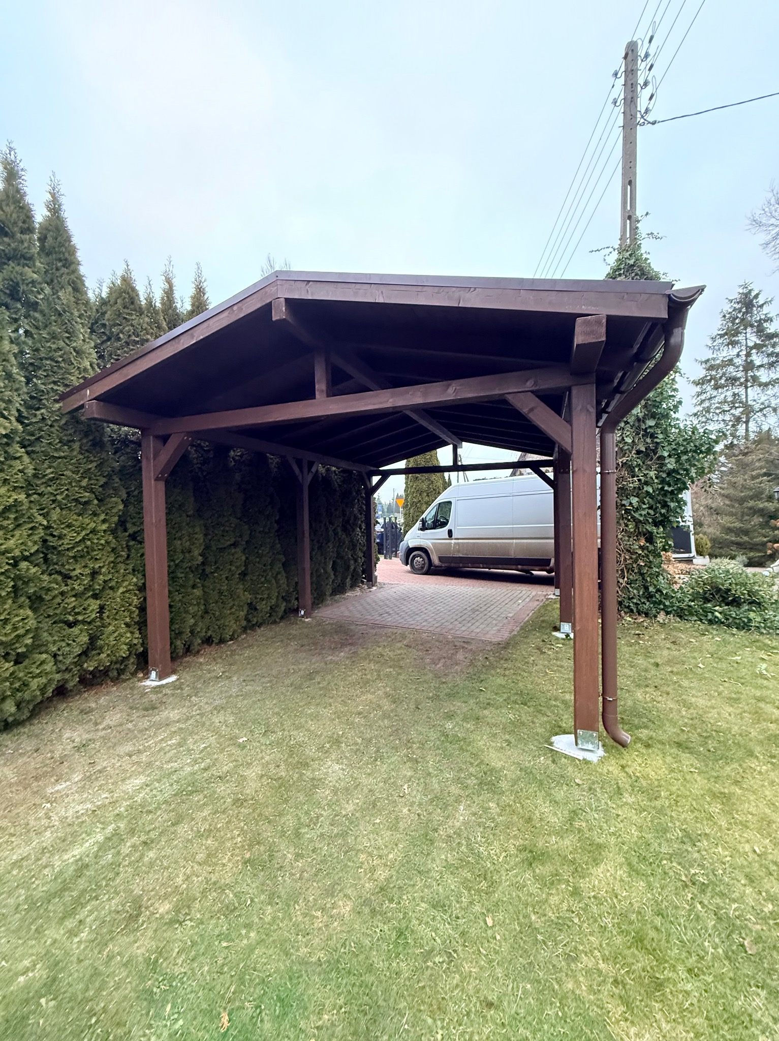 Ciemnobrązowa, drewniana wiata garażowa na trawniku, z zaparkowanym białym vanem w tle. Konstrukcja z rynną, na betonowej podstawie. Otoczenie: tuje, drzewa.