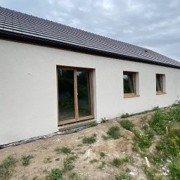 Vladyslav Krasovskyi ProFundation - Jasna elewacja domu jednorodzinnego z brązowymi oknami i ciemnym dachem, widok z boku na tle pochmurnego nieba i ziemi z roślinnością.