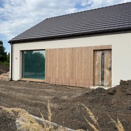 Vladyslav Krasovskyi ProFundation - Nowoczesny dom jednorodzinny z drewnianą elewacją i dużym oknem, częściowo zakrytym folią ochronną. Widoczne prace ziemne wokół budynku i brązowy dach.