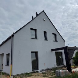 Vladyslav Krasovskyi ProFundation - Dom jednorodzinny z szarą elewacją i czarnymi oknami, widoczny z dołu pod kątem. W tle pochmurne niebo. Częściowo widoczna konstrukcja z czarną membraną.