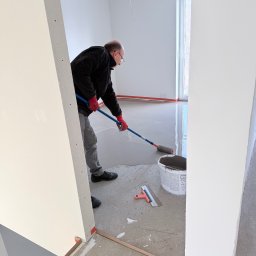 Vladyslav Krasovskyi ProFundation - Wylewka samopoziomująca rozprowadzana wałkiem w jasnym pomieszczeniu. Widoczny pracownik w czerwonych rękawicach, wiadro z masą i szpachelka. Podłoże zabezpieczone taśmą.