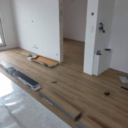 Vladyslav Krasovskyi ProFundation - Układanie paneli podłogowych w jasnym wnętrzu. Widoczne rozłożone panele, folia izolacyjna, kartony i narzędzia. Praca w toku, częściowo ułożona podłoga.