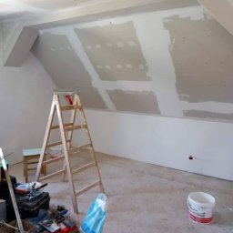 Vladyslav Krasovskyi ProFundation - Poddasze w trakcie wykańczania: widoczna drewniana drabina, narzędzia, worki z gipsem i częściowo wykończone ściany z płyt kartonowo-gipsowych.