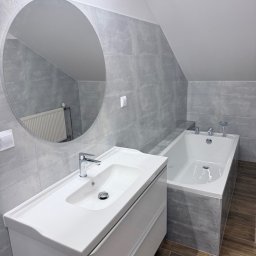 Vladyslav Krasovskyi ProFundation - Wykończona łazienka na poddaszu: szare płytki imitujące kamień, biała umywalka z szafką i okrągłe lustro. W odbiciu lustra widoczny grzejnik.