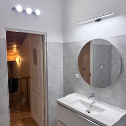 Vladyslav Krasovskyi ProFundation - Wykończona łazienka z szarymi płytkami, okrągłym lustrem i białą szafką pod umywalką. Widoczne oświetlenie LED i otwarte drzwi do innego pomieszczenia z drewnianym wykończeniem.