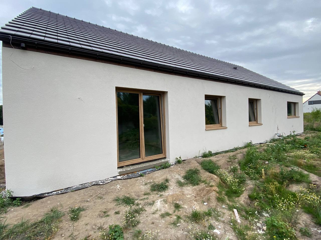 Jasna elewacja domu jednorodzinnego z brązowymi oknami i ciemnym dachem, widok z boku na tle pochmurnego nieba i ziemi z roślinnością.