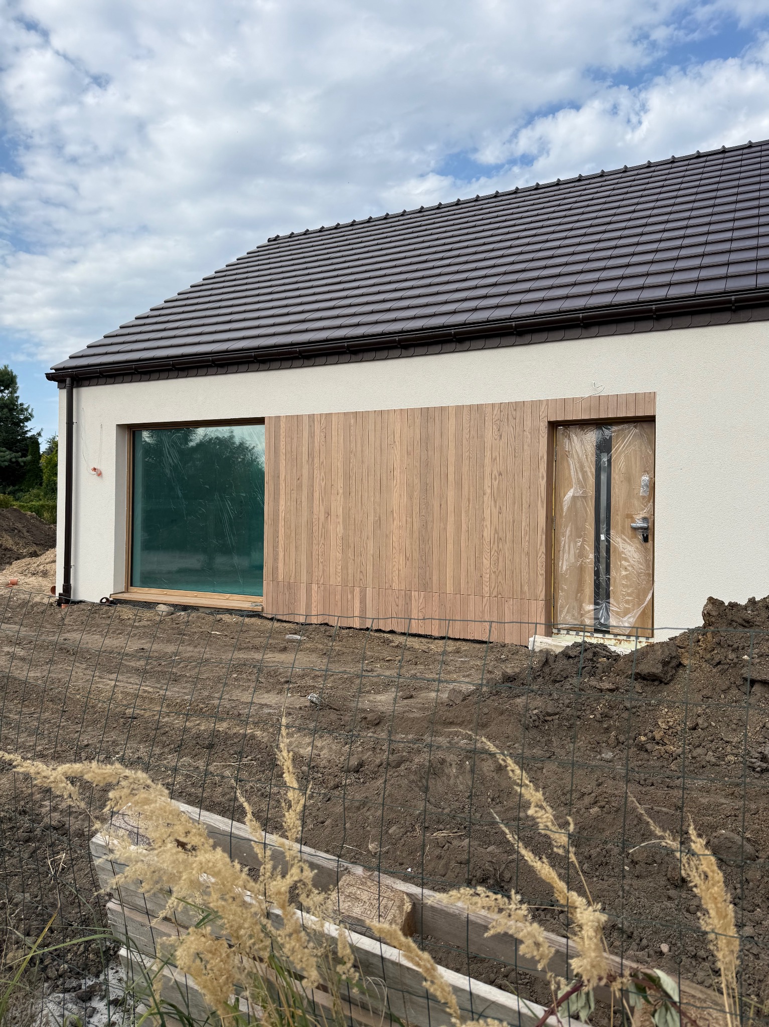 Nowoczesny dom jednorodzinny z drewnianą elewacją i dużym oknem, częściowo zakrytym folią ochronną. Widoczne prace ziemne wokół budynku i brązowy dach.