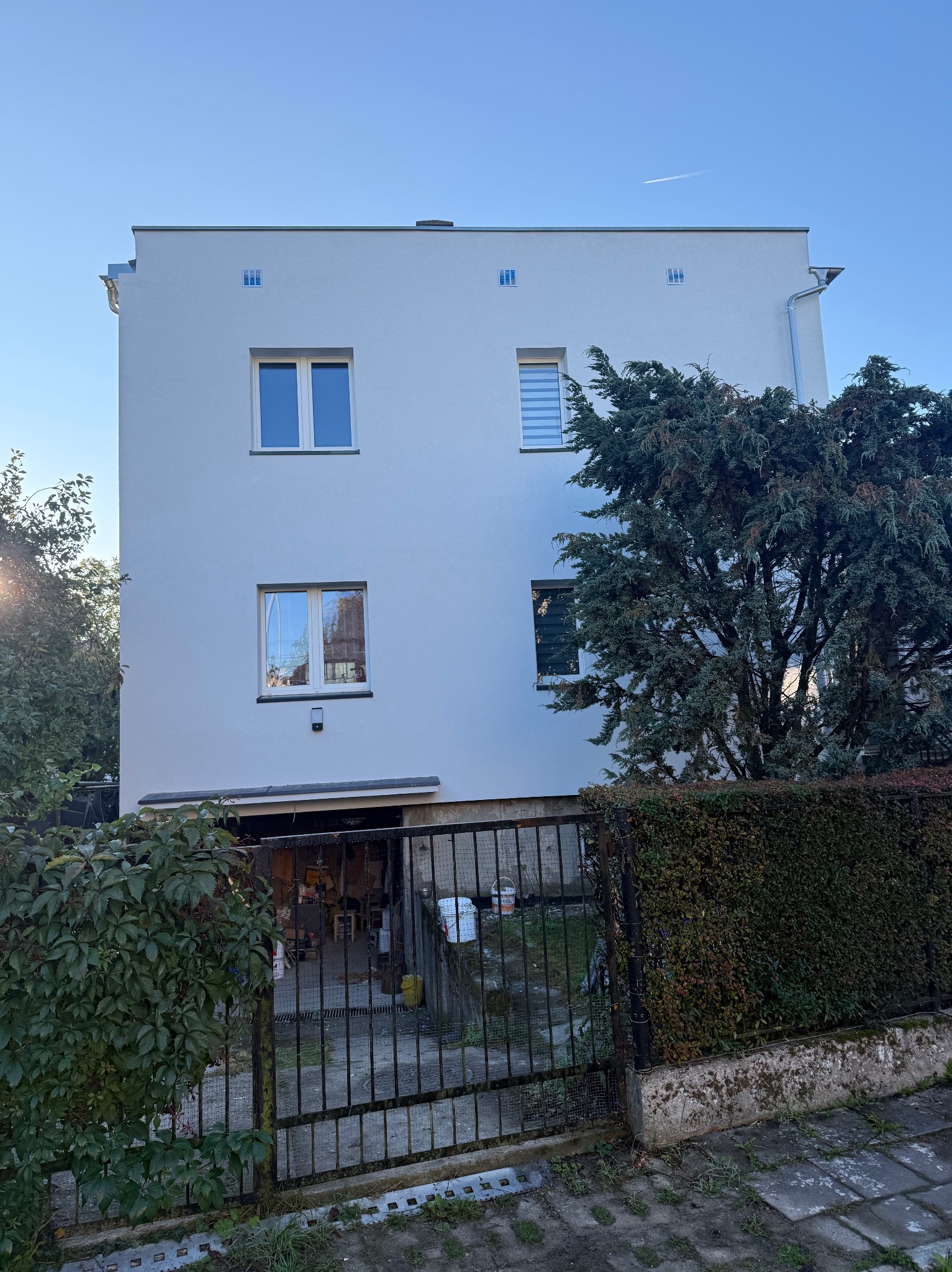 Biała, odnowiona elewacja trzypiętrowego budynku z widocznymi oknami i zielenią. Ujęcie z poziomu ulicy, z metalowym ogrodzeniem na pierwszym planie.