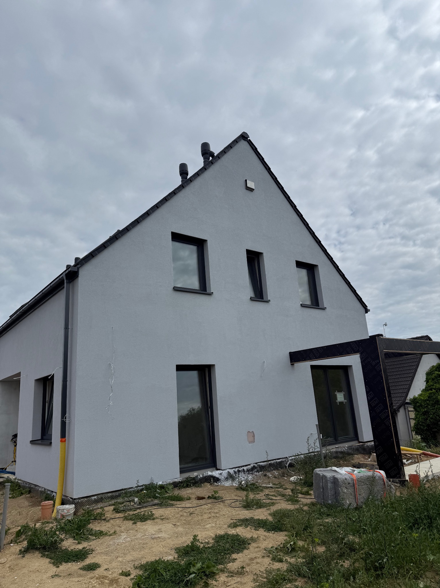 Dom jednorodzinny z szarą elewacją i czarnymi oknami, widoczny z dołu pod kątem. W tle pochmurne niebo. Częściowo widoczna konstrukcja z czarną membraną.