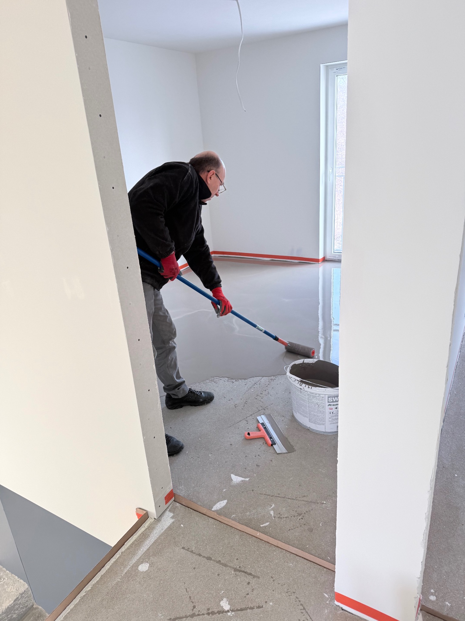 Wylewka samopoziomująca rozprowadzana wałkiem w jasnym pomieszczeniu. Widoczny pracownik w czerwonych rękawicach, wiadro z masą i szpachelka. Podłoże zabezpieczone taśmą.