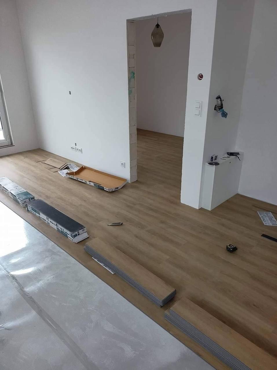 Układanie paneli podłogowych w jasnym wnętrzu. Widoczne rozłożone panele, folia izolacyjna, kartony i narzędzia. Praca w toku, częściowo ułożona podłoga.