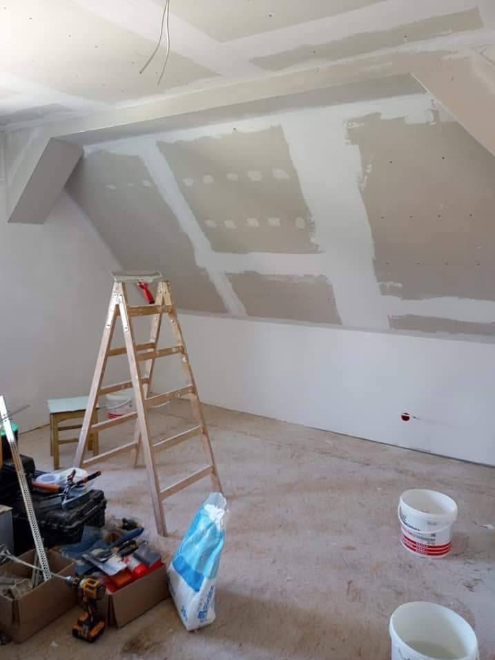 Poddasze w trakcie wykańczania: widoczna drewniana drabina, narzędzia, worki z gipsem i częściowo wykończone ściany z płyt kartonowo-gipsowych.