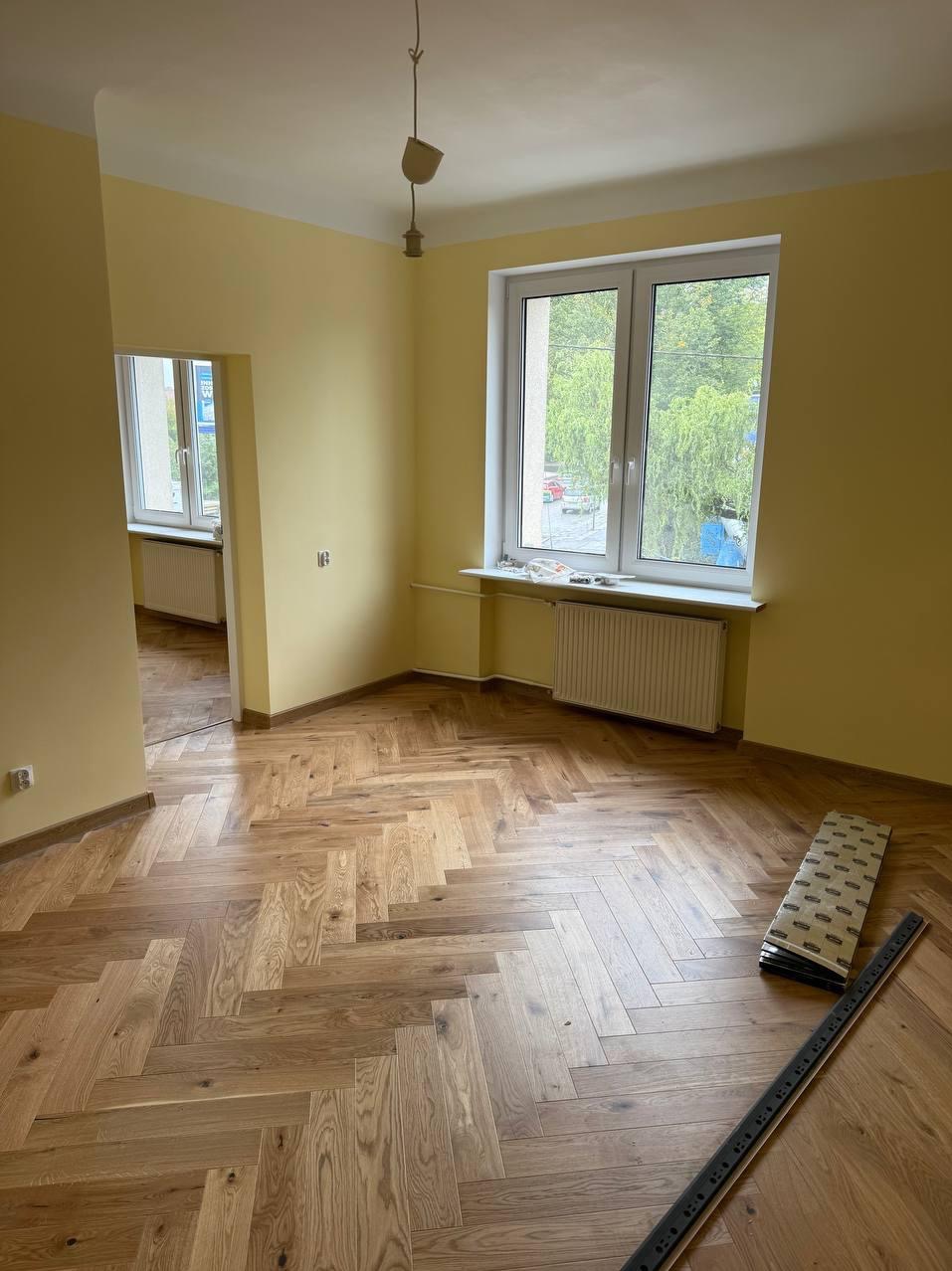 Parkiet ułożony w jodełkę w pokoju z żółtymi ścianami. Widoczne listwy przypodłogowe i grzejnik pod oknem. Częściowo widoczne panele i listwa pomiarowa.