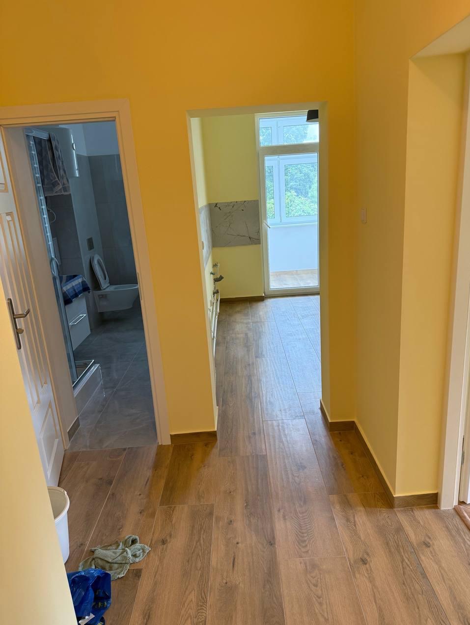 Jasne wnętrze po remoncie: drewnopodobna podłoga, żółte ściany, widok na łazienkę z kabiną prysznicową i toaletą oraz kuchnię z balkonem. Perspektywa z korytarza.