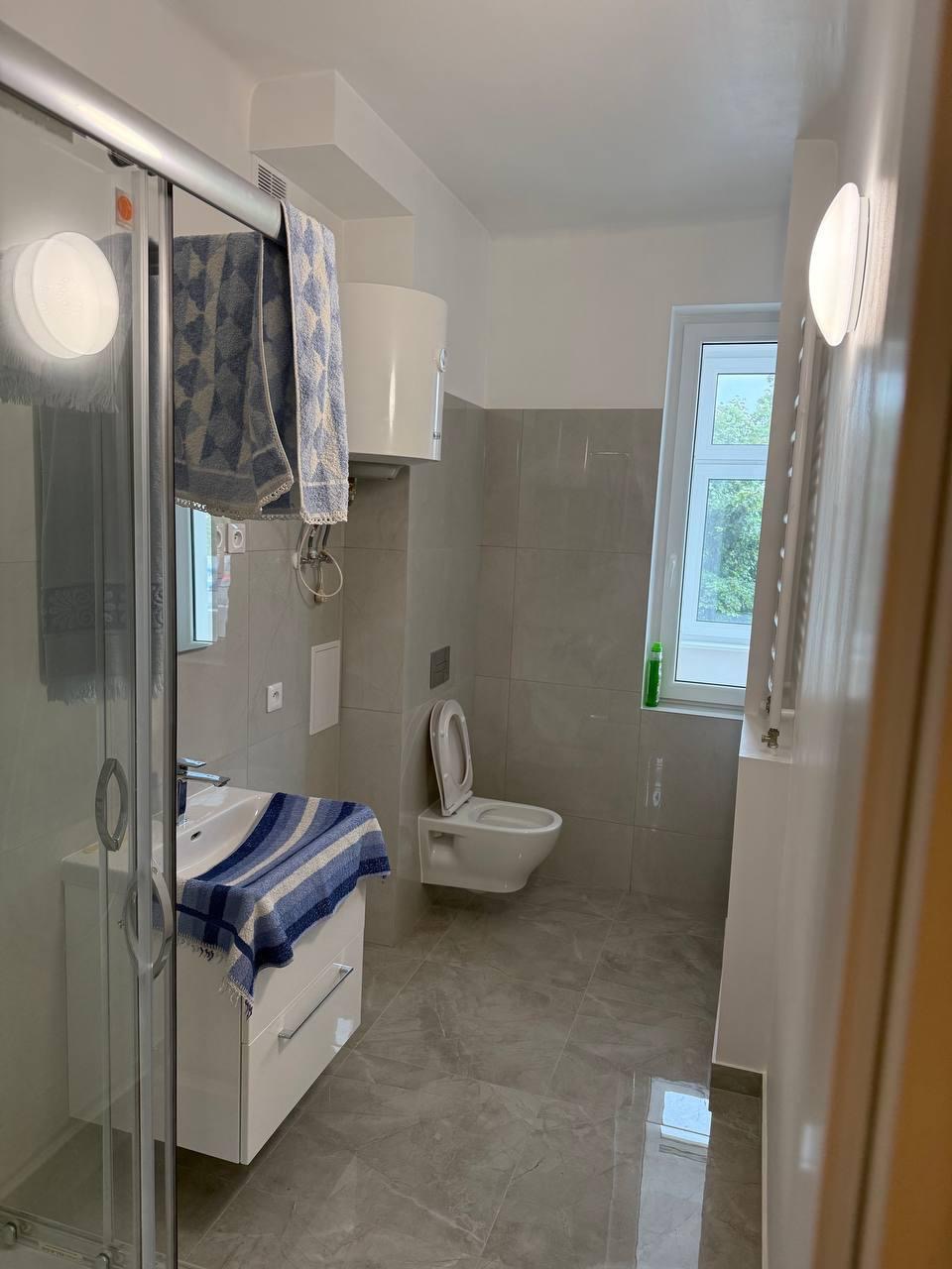 Jasna łazienka z szarymi płytkami, białą armaturą i szafką pod umywalką. Widoczny fragment kabiny prysznicowej i okno z zielenią za szybą. Minimalistyczny styl.