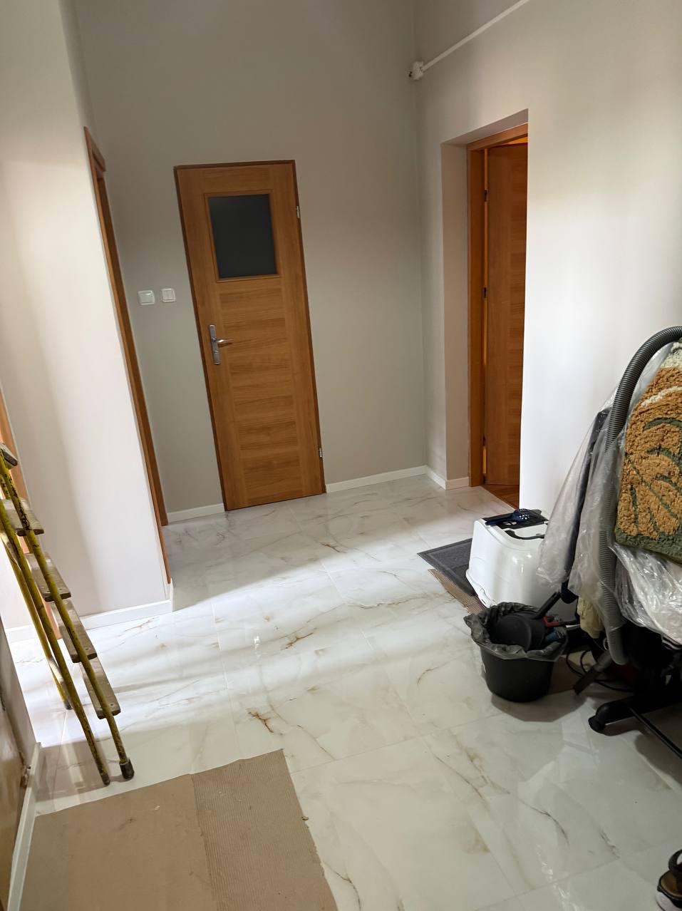 Jasne wnętrze po remoncie: białe marmurowe płytki podłogowe, świeżo pomalowane ściany w odcieniach beżu, drewniane drzwi z szybą. Widoczna drabina i sprzęt sprzątający.